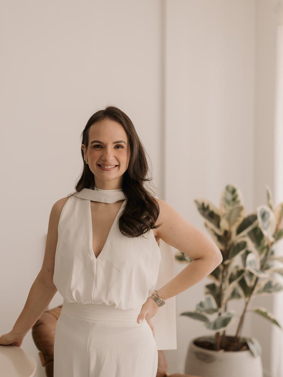 Beatriz Aires, de macacão branco, em pé e sorrindo, com a mão na cintura