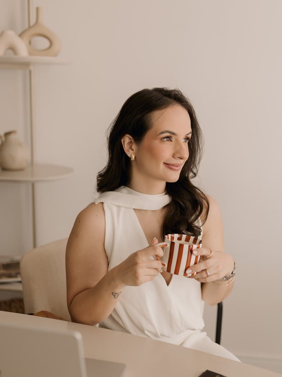 Beatriz Aires, de branco, segura uma caneca de café à mesa de trabalho e sorri