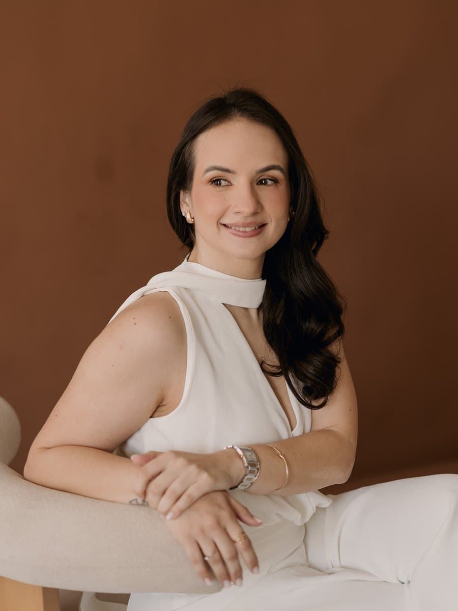 Beatriz Aires, de branco, sentada em uma poltrona clara diante de um fundo marrom, sorrindo